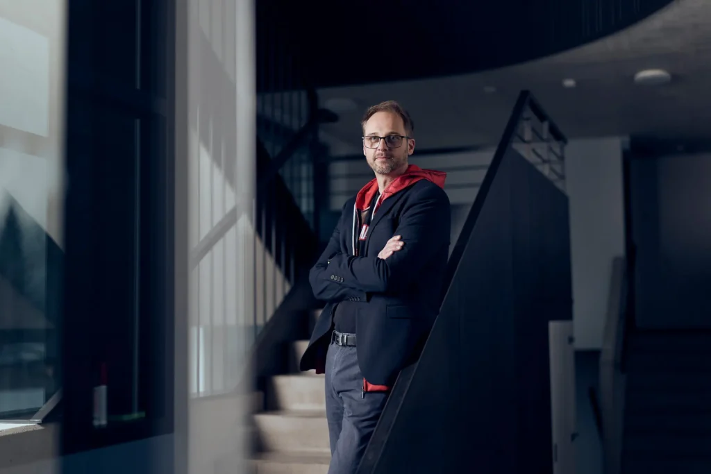 Business‑Porträt einer Person in professioneller Kleidung vor moderner, geometrischer Treppenarchitektur in einem Bürogebäude.